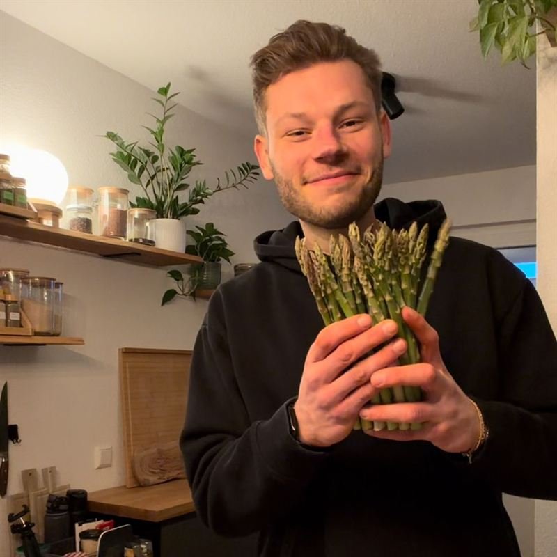 Max steht lachend in seiner Küche und hält einen Bund grünen Spargel in den Händen